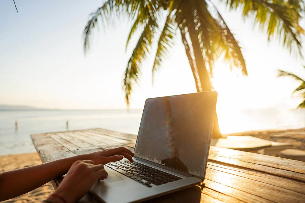 Someone working a laptop on a table beside a beach